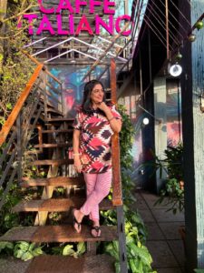 A photograph of Abha Mehta standing on the stairs on a cafe, with a lot of greenery around her 