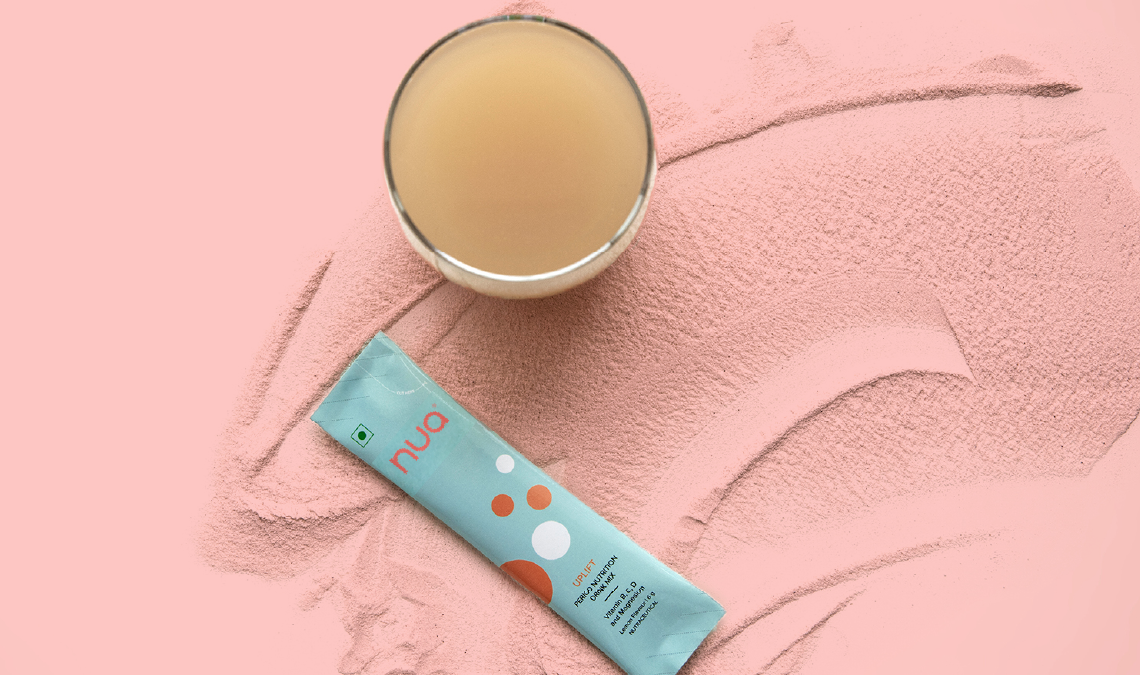 A glass of beige-colored drink placed on a surface dusted with pink powder, alongside a sachet of Nua Uplift pain-relief drink mix.