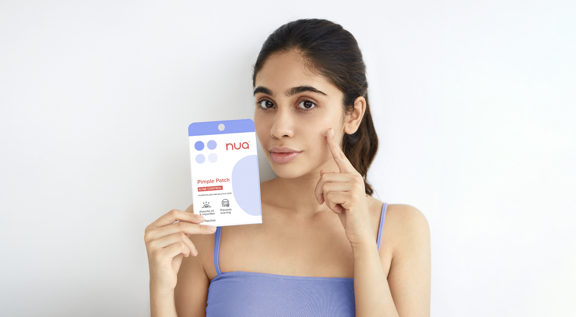 Young woman in a blue top holding a pack of Nua Pimple Patch while pointing to her cheek, promoting acne care.