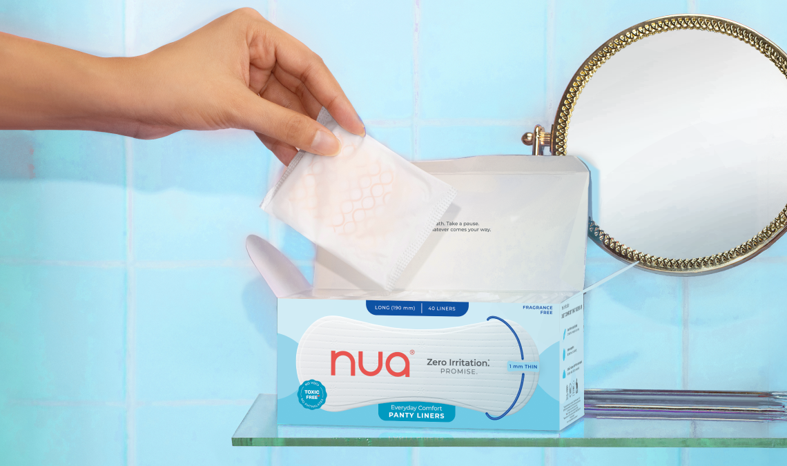 A hand pulling out a panty liner from an open box of Nua Everyday Comfort Panty Liners, placed on a glass shelf in a bathroom with a blue tiled wall and a round mirror in the background.