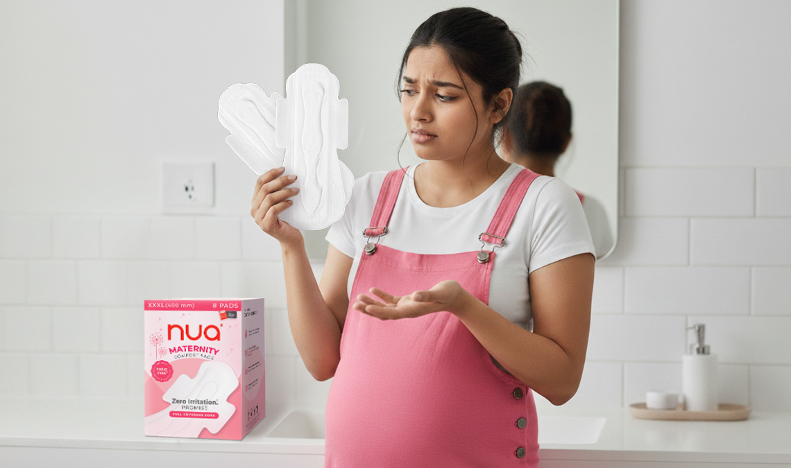 Pregnant woman in pink overalls holding multiple maternity pads with a confused expression, standing in a bathroom beside a box of Nua Maternity Comfort Pads.