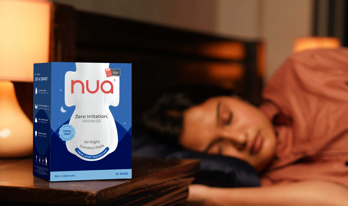 A box of Nua All Night Comfort Pads placed on a bedside table next to a lit lamp, with a person sleeping peacefully in the background, suggesting overnight protection.