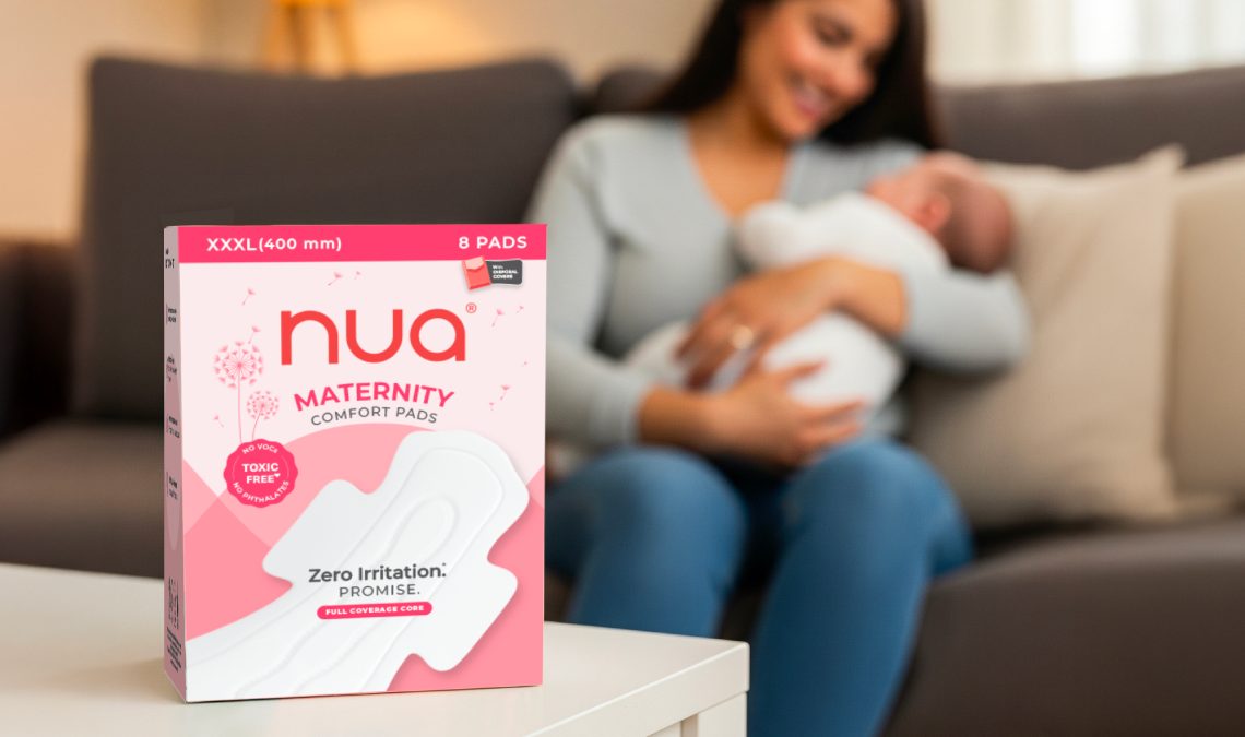 A box of Nua Maternity Comfort Pads placed on a white table, with a smiling mother in the background sitting on a couch and holding her baby, highlighting the product’s use for postpartum care.