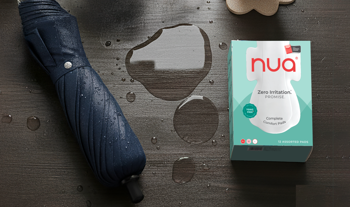 A box of Nua Complete Comfort Pads placed on a wet wooden surface with water droplets and a closed navy umbrella beside it, suggesting monsoon season.