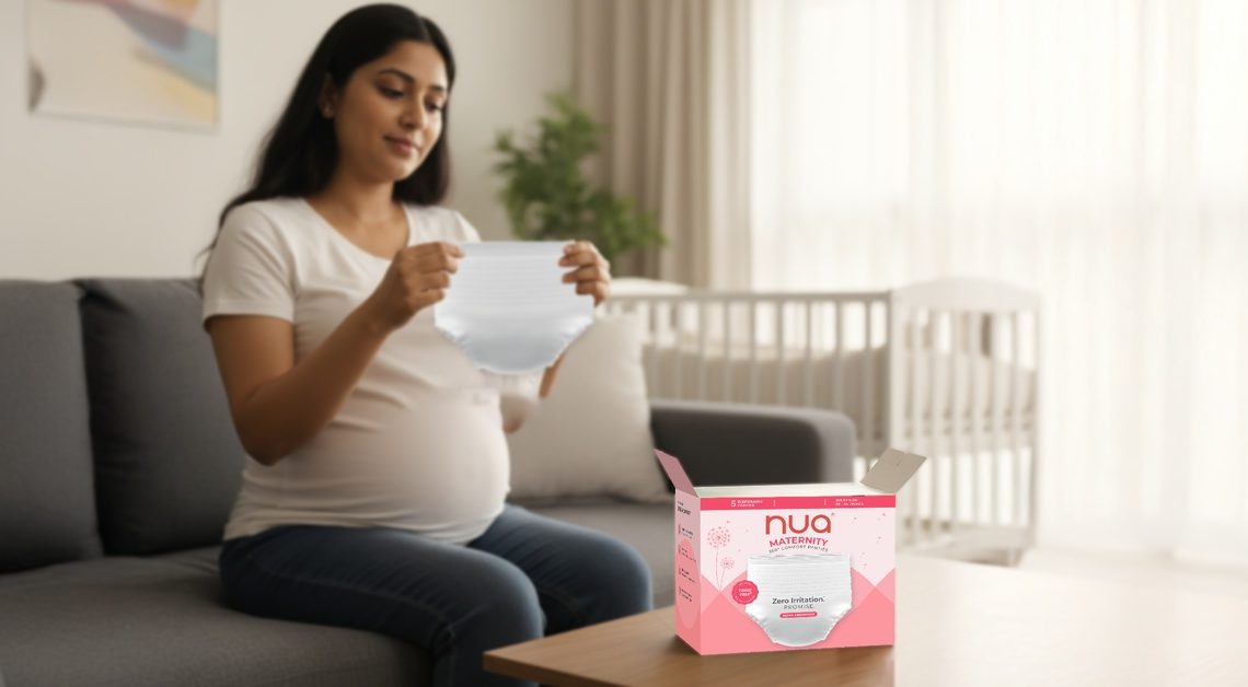Pregnant woman sitting on a couch, smiling while holding Nua maternity panties, with a box of maternity panties on the table beside her.