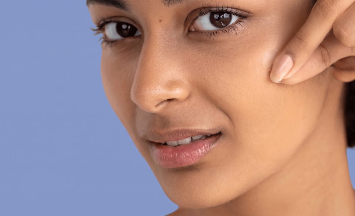 Close-up of a person applying a small pimple patch to their cheek with a fingertip against a soft blue background.